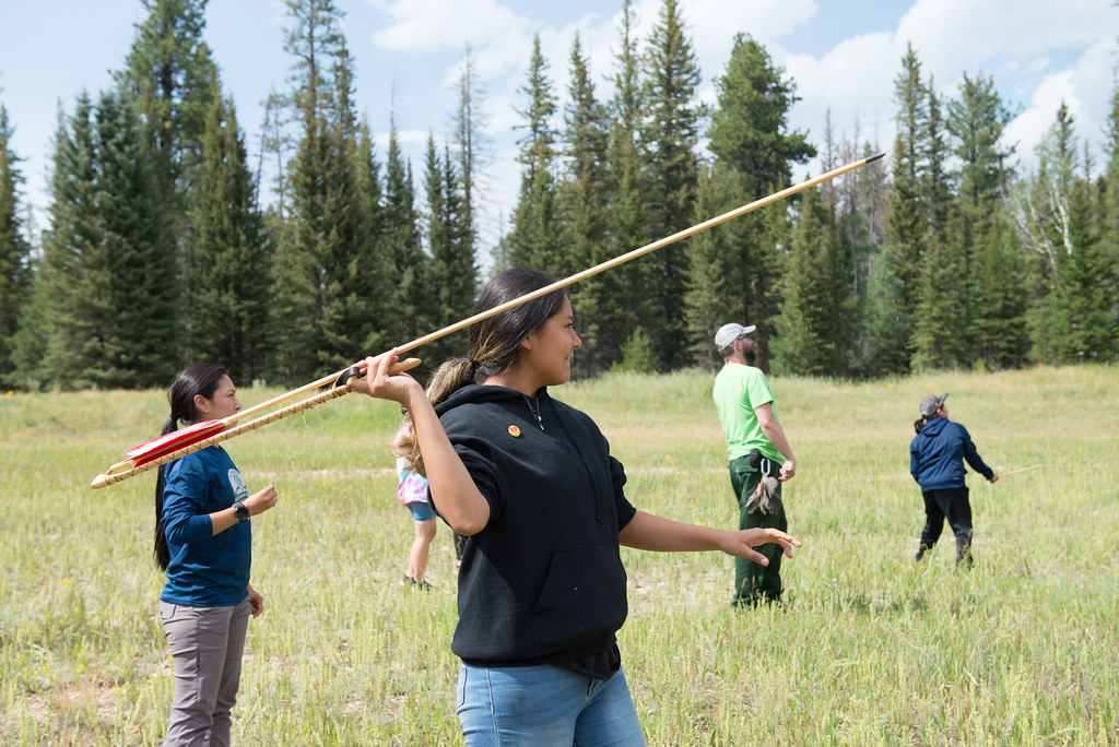 A girl standing in a field surrounded by trees uses an atlatl to throw a long arrow.