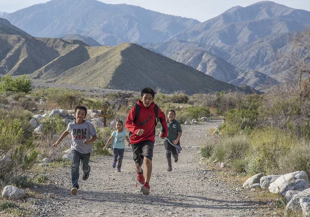 Four young children running down a gravel path, with hills in the background and dry scrubby grass around them