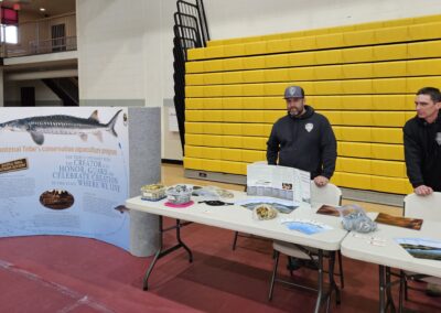 Two men stand behind a table with informational brochures. Next to the table is a sign with a sturgeon describing conservation aquaculture.