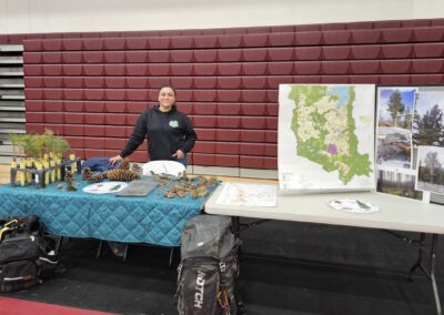 Woman stands behind table with pine cones and pine saplings - to one side there is another table with a map of the Flathead reservation and pictures of tree conservation work